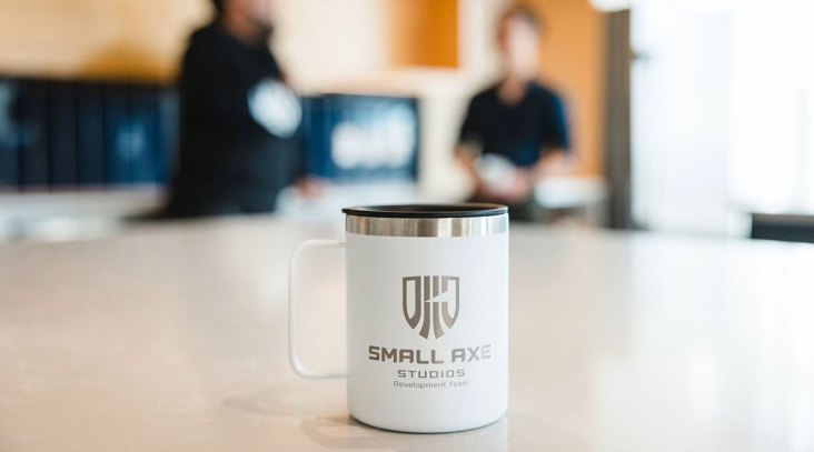 نمای نزدیک از ماگ سفید اختصاصی با لوگوی حکشده Small Axe Studios در محیط داخلی دفتر بازیسازی